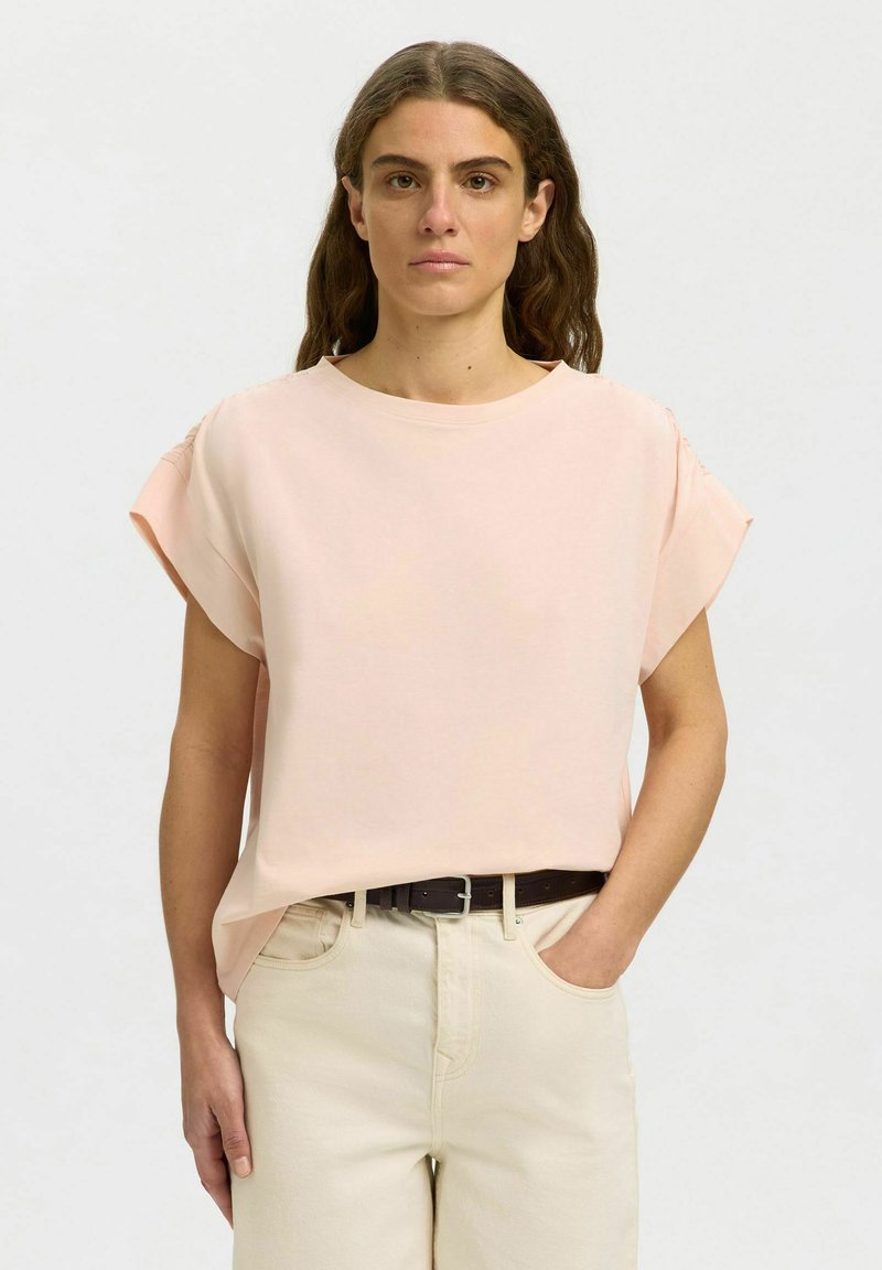 Femme aux cheveux bruns ondulés portant un chemisier rose pâle à manches courtes et un pantalon beige clair, debout avec une main dans la poche.