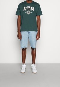 Hombre con camiseta oscura verde de Adidas, pantalones cortos de mezclilla claros y desgastados, y zapatillas altas blancas de Adidas, apoyado contra una pared blanca.