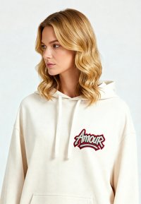 Felpa color crema realizzata in morbido tessuto, caratterizzata da un grande logo ricamato "Amour" in rosso e grigio. Include coulisse e una tasca frontale.