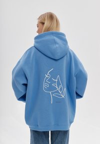 Felpa blu oversize con cappuccio regolabile, caratterizzata da un disegno a linee bianche di un volto e foglie sul retro. Realizzata in cotone.