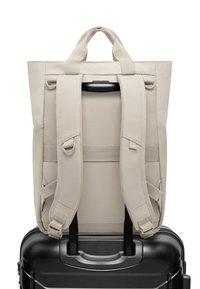 Beige hátizsák állítható pántokkal és felső fogantyúval, fekete kerekes bőröndön pihen. Tartós anyagból készült, minimalista design jellemzi.