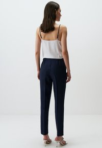 Navy kleurige broek met een relaxte pasvorm en een elastische tailleband, gecombineerd met een witte top met dunne bandjes. Achtergrond is eenvoudig.