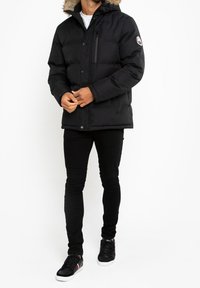 Chaqueta de plumas negra con capucha forrada de piel sintética desmontable, bolsillo en el pecho con cremallera y cierre de botones a presión, combinada con pantalones negros ajustados y zapatillas.