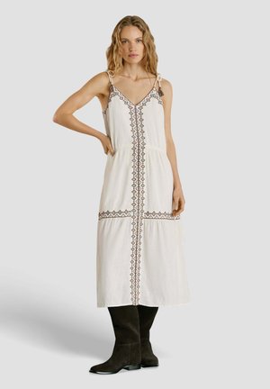 Frau trägt ein weißes ärmelloses Midikleid mit geometrischer Stickerei, gebundenen Schulterriemen und schwarzen kniehohen Stiefeln, steht mit einer Hand in der Hüfte.