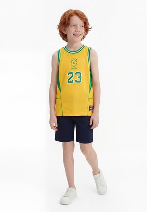 Ένα αγόρι με κόκκινα μαλλιά χαμογελά, φοράει κίτρινη μπλούζα μπάσκετ της Βραζιλίας με τον αριθμό 23, ναυτικό σορτς και λευκά αθλητικά παπούτσια, στέκεται σε λευκό φόντο.