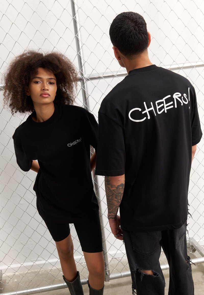 Svart oversized t-skjorter med "CHEERS!" i hvit tekst på front og bak. Modell iført svarte sykkelshorts og støvler, mot en kjettinglinkbakgrunn.