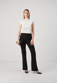 Hvit ermeløs topp med høy krage, kombinert med svarte skreddersydde bukser og svarte spisse hæler, holder en svart clutch.