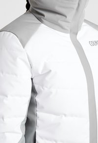 Puffjacka i vitt med grå accenter, med hög krage, kviltad textur och synlig sömdetalj. Varumärkeslogotyp på sidan.