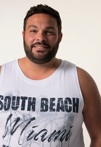 Bílý top bez rukávů s černou grafikou "SOUTH BEACH Miami", kulatým výstřihem a měkkou bavlněnou texturou. Model má vousy a krátké vlasy.