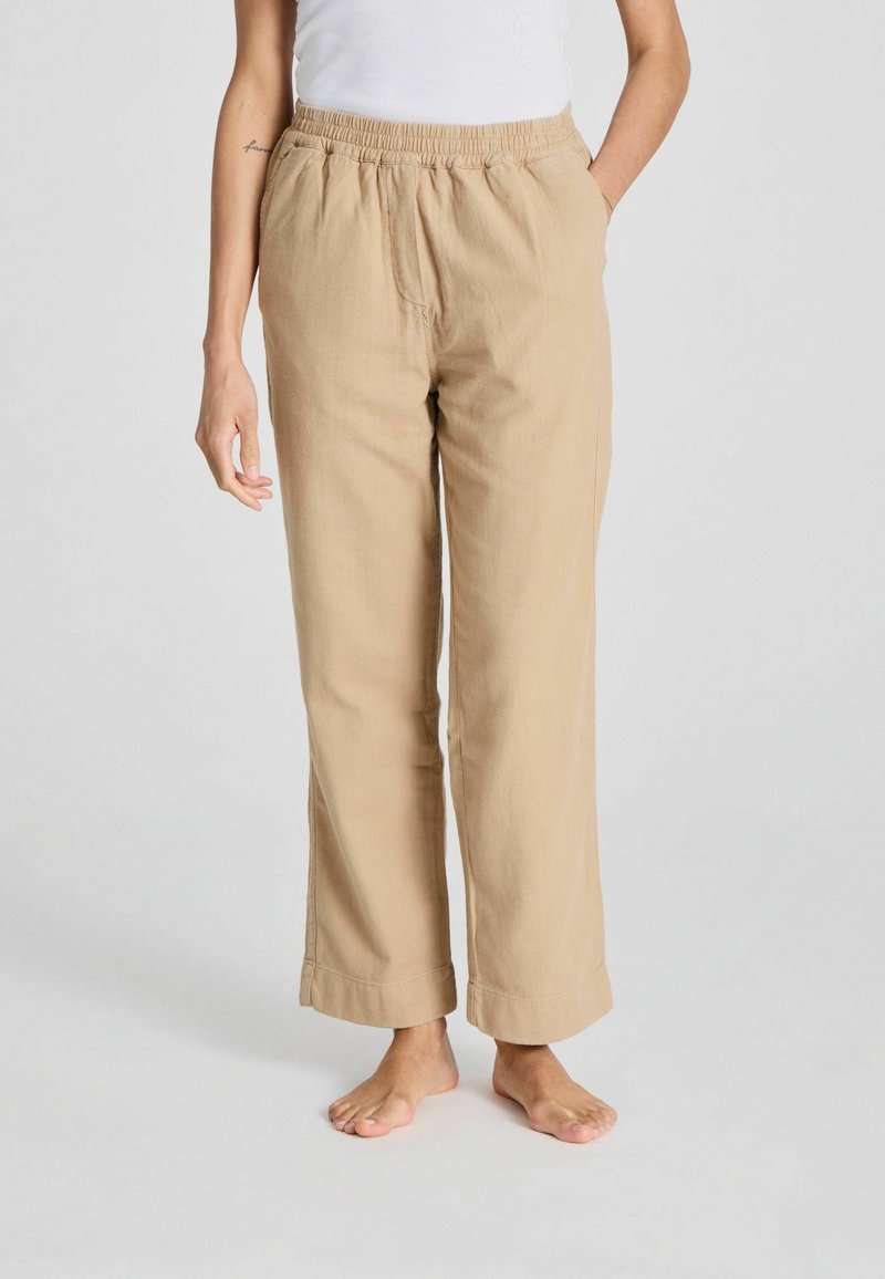 Pantalones de lino beige con cinturilla elástica, dos bolsillos laterales y piernas anchas. La textura parece ligera y transpirable.