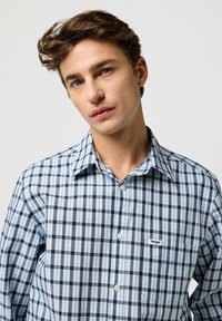 Camicia a quadri blu e bianca con chiusura a bottoni, dotata di taschino sul petto e logo "Wrangler". Realizzata in tessuto leggero.