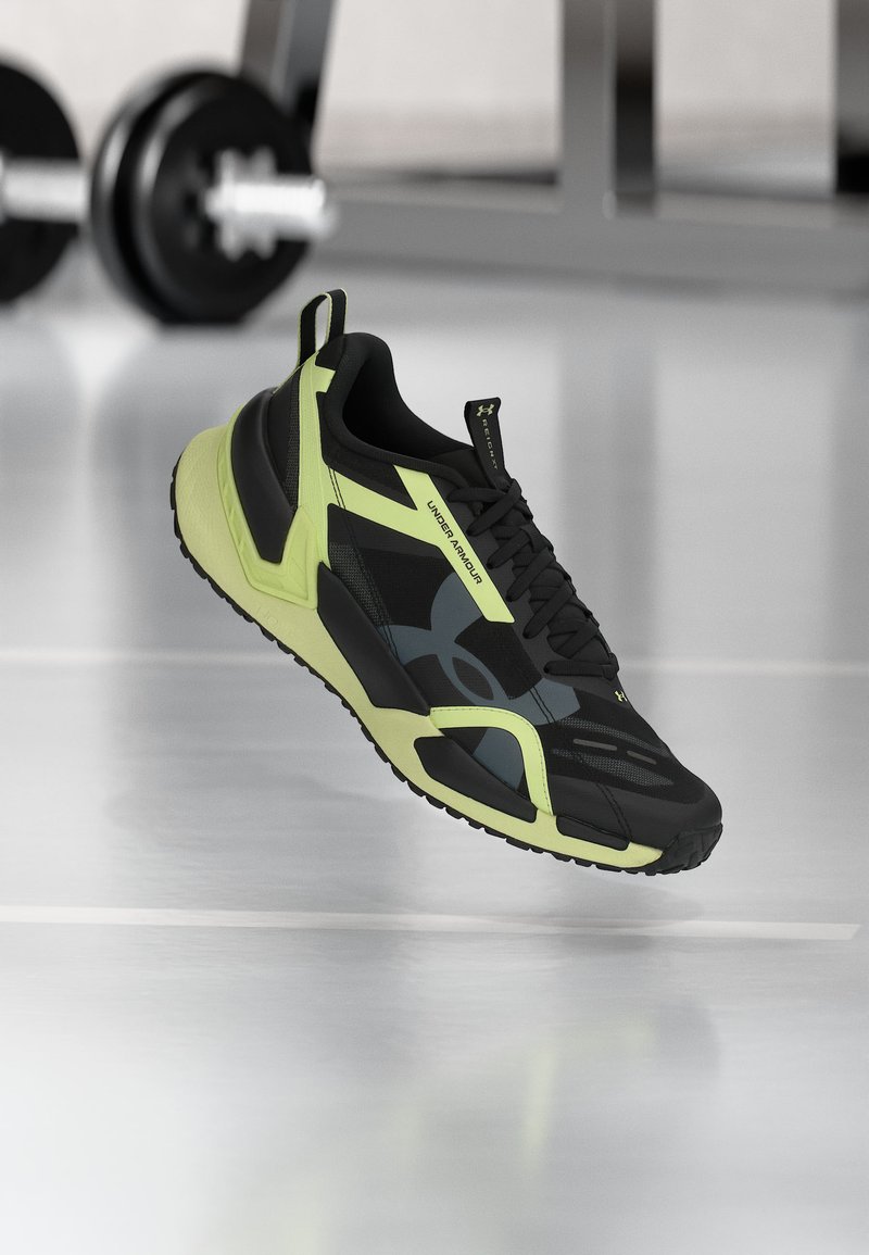 Μαύρο και νέον πράσινο αθλητικό παπούτσι Under Armour που αιωρείται σε ένα γυμναστήριο με μια μπάρα βάρη στο φόντο.