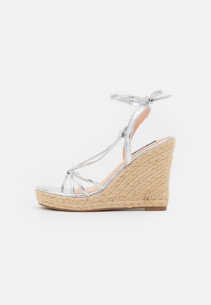 Sølvfarvet kilehæl sandal med remme og ankelbånd samt flettet hæl i naturfiber, vist fra siden på hvid baggrund.