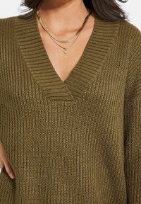 Olivgrüner Strickpullover mit V-Ausschnitt und gerippter Textur. Verfügt über geschichtete goldene Halsketten mit einem runden Anhänger.
