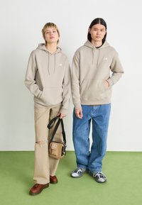 Le felpe beige presentano tasche a marsupio e cordini. Un modello indossa pantaloni chiari strappati e scarpe marroni, mentre l'altro indossa jeans blu e sneakers.