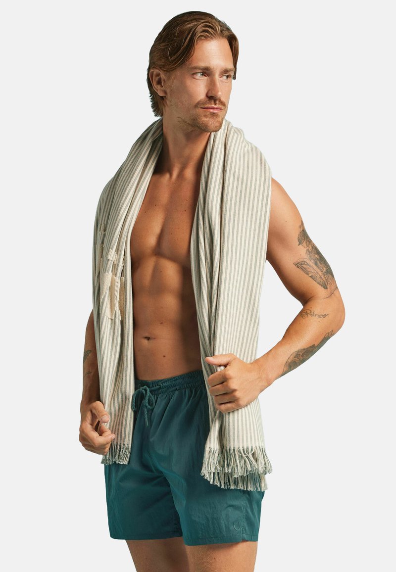Serviette en coton rayée beige et crème, portée sur un torse nu. Le modèle porte également un maillot de bain teal avec une taille élastique et un cordon de serrage.