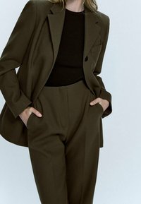 Costume vert olive foncé comprenant un blazer ajusté avec un bouton unique et un pantalon taille haute assorti, associé à un top noir moulant.