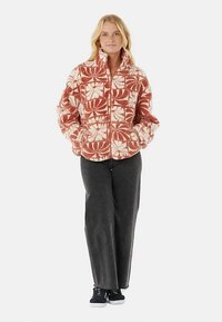 Fleece jacket with a red and white floral pattern, zippered front, high collar, and two side pockets. Paired with dark gray pants and sneakers.