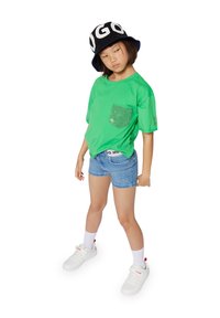 Camiseta verde de manga corta con un bolsillo de lentejuelas, combinada con pantalones cortos de mezclilla azul claro y zapatillas blancas. Sombrero bucket negro con texto blanco.