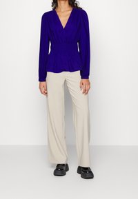 Blusa a maniche lunghe a scollo a V in blu reale con spalle arricciate, vita cinte e tessuto morbido; abbinata a pantaloni larghi beige e scarpe nere.
