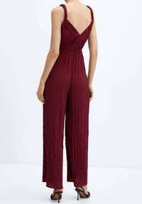 Combinaison bordeaux en tissu texturé plissé ; coupe jambes larges ; dos en forme de V ; bretelles épaisses ; associée à des sandales noires à talons hauts.