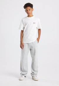 Valkoinen puuvillapaita lyhyillä hihoilla ja pienellä punaisella logolla, yhdistettynä vaaleanharmaisiin collegehousuihin ja valkoisiin lenkkareihin.