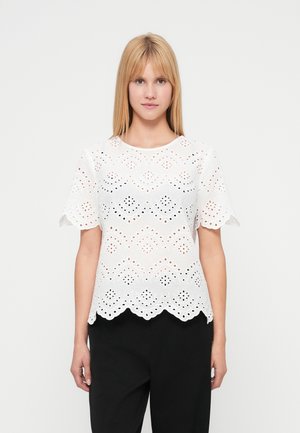 Λευκό μπλουζάκι με διακοσμητικές τρυπούλες, με κυματιστό τελείωμα, κοντά μανίκια και περίπλοκα μοτίβα με κοψίματα. Φορεμένο πάνω από μαύρο παντελόνι.