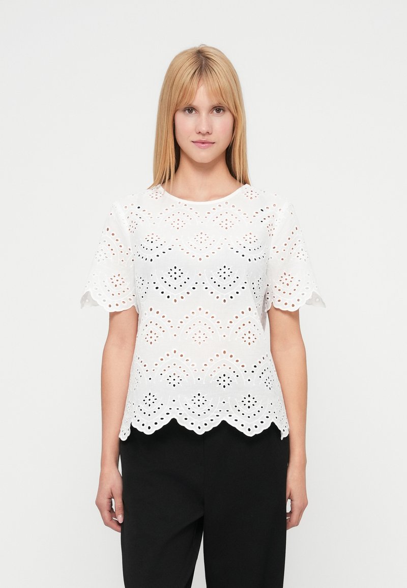 Λευκό μπλουζάκι με διακοσμητικές τρυπούλες, με κυματιστό τελείωμα, κοντά μανίκια και περίπλοκα μοτίβα με κοψίματα. Φορεμένο πάνω από μαύρο παντελόνι.