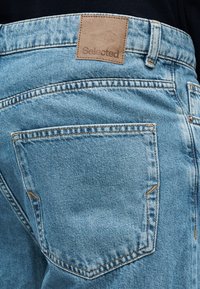 Hele sinised teksapüksid, mille püksirihmal on nahast silt "Selected". Tagataskutel on tavaline oranži õmblusega viimistlus.