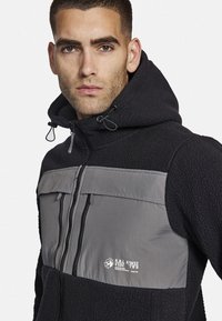 Chaqueta de forro polar con un torso de color azul marino oscuro y un panel gris en el pecho. Presenta un cierre frontal con cremallera y una capucha ajustable con cordones.