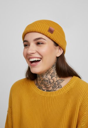 Mustard yellow knit beanie with ribbed texture and a small leather patch. Worn with a matching knitted sweater featuring a round neckline.