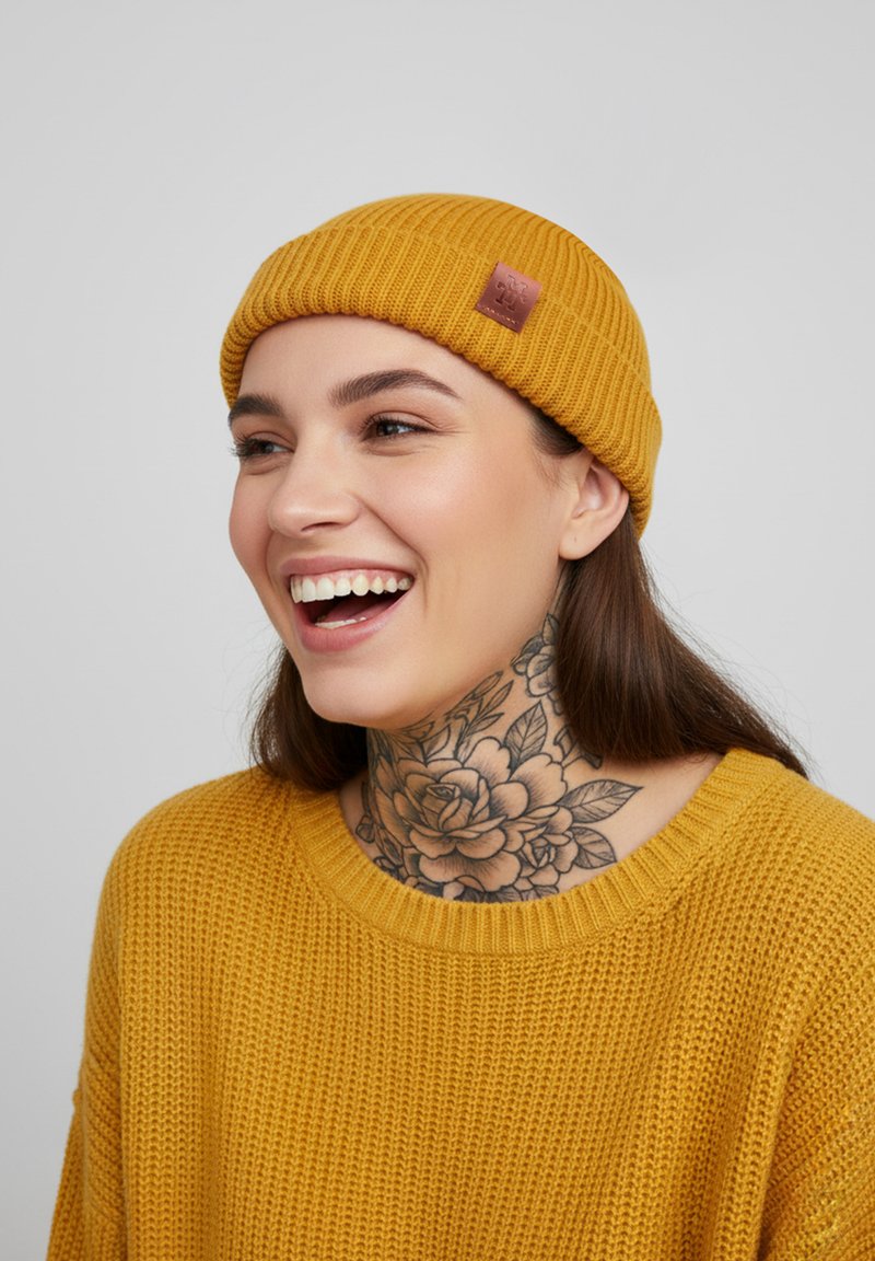 Mustard yellow knit beanie with ribbed texture and a small leather patch. Worn with a matching knitted sweater featuring a round neckline.