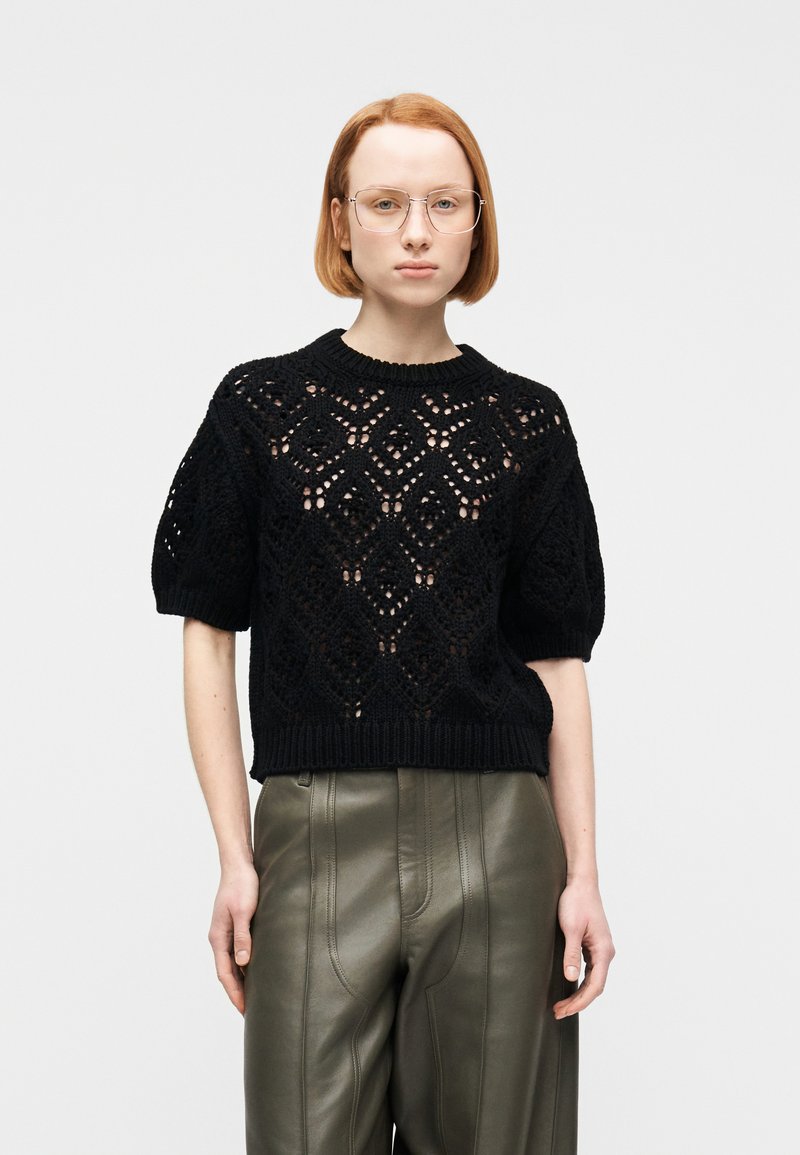 Tânără purtând ochelari transparenți, un top negru tricotat cu mâneci scurte și modele în formă de diamant, și pantaloni din piele verde măsliniu, cu talie înaltă.