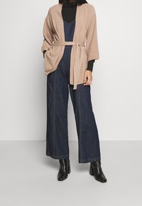 Beige gebreide cardigan met brede mouwen, vastgebonden in de taille, over een donkerdenim jumpsuit. Zwarte hooggehakte laarzen maken de look compleet.