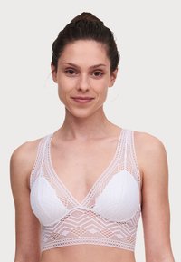 Bralette bianco in pizzo con un motivo geometrico, scollatura profonda, spalline larghe e una finitura morbida e testurizzata. Presenta dettagli in pizzo trasparente.