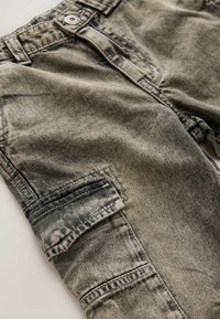 Pantaloni cargo in denim verde oliva sbiadito, caratterizzati da una tasca laterale, cuciture visibili e una leggera variazione di texture sul tessuto.