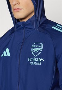 Giacca sportiva blu navy con cappuccio, caratterizzata dal logo dell'Arsenal in azzurro chiaro e dal testo "Emirates Fly Better". Realizzata in materiale leggero e liscio.