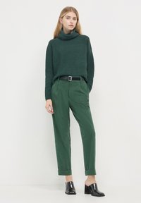 Dunkelgrüner, gerippter Rollkragenpullover, kombiniert mit passenden grünen Cordhosen, ausgestattet mit einem schwarzen Gürtel und glatten schwarzen Absatzmules.