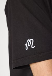 Juodas marškinėliai su balta stilizuota logotipu ant rankovės. Pagaminti iš minkštos medžiagos, drabužiai turi trumpas rankoves ir apvalų kaklą.