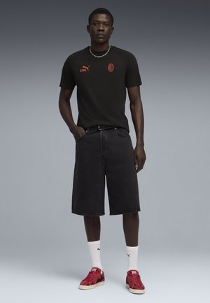 Jeune homme debout portant un t-shirt noir Puma avec un logo rouge, un short long noir, des chaussettes blanches et des baskets rouges Puma sur fond gris.