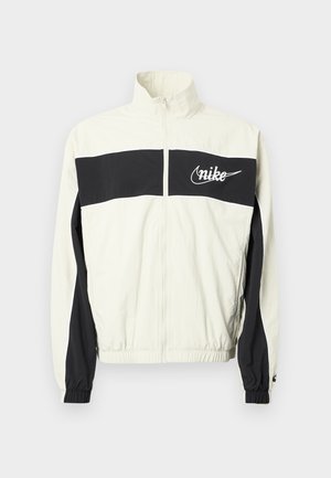 Vit och svart Nike-jacka med hög krage, dragkedja framtill och elastiska muddar. Har en tydlig svart rand över bröstet.