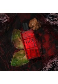 Bouteille en verre rouge rectangulaire étiquetée "DAVIDOFF HOT WATER", située sur des feuilles vertes avec des gouttes, sur un fond sombre et rocheux.