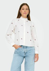 Donna dai lunghi capelli che indossa una blusa bianca con piccoli motivi floreali rossi e jeans azzurri chiari, con la mano in tasca, su uno sfondo neutro.