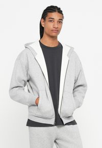 Fiatal személy, aki fonott hajat visel, szürke zipzáros kapucnis pulóvert és fekete pólót, valamint azonos színű szürke melegítőt visel, keze a zsebében áll.