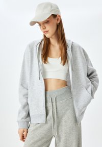 Felpa con zip grigia chiara, dal fit rilassato e con tasche, abbinata a un crop top bianco e pantaloni della tuta grigi abbinati, con cordini.