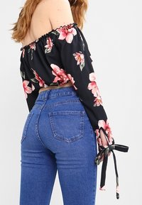 Svart off-shoulder topp med blommönster, rynkad halsringning och knytdetaljer vid ärmarna, kombinerad med högmidjade blå denimjeans.