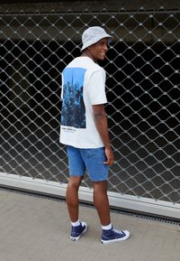 Witte t-shirt met korte mouwen met een blauwe stadsafbeelding op de achterkant, gecombineerd met een blauwe denimshort en marineblauwe hoge sneakers.