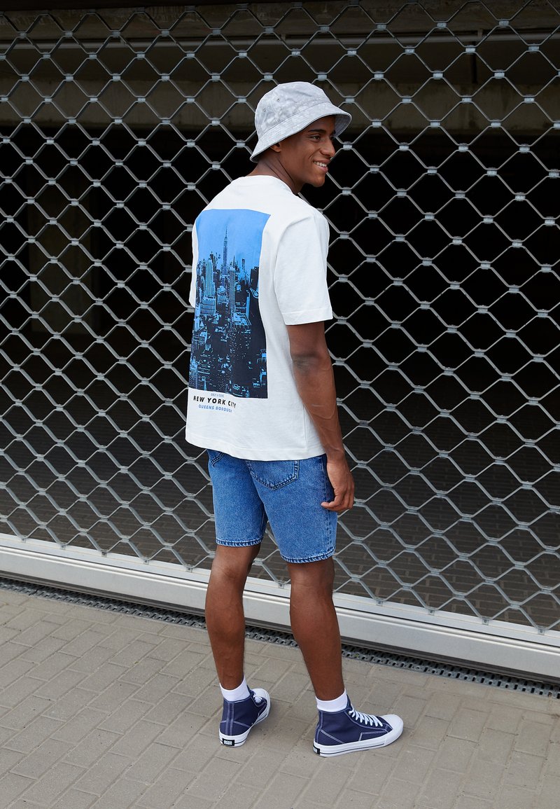 Witte t-shirt met korte mouwen met een blauwe stadsafbeelding op de achterkant, gecombineerd met een blauwe denimshort en marineblauwe hoge sneakers.