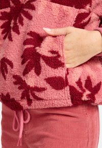 Flauschiger Fleece-Oberteil in Rosa mit burgunderfarbenem Blumenmuster, ausgestattet mit einer Fronttasche und einem Kordelzug am Saum. Kombiniert mit rosa Cordhose.