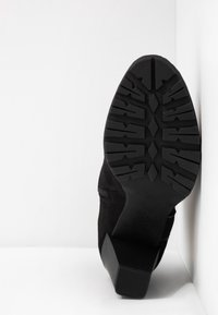 Botín de ante negro con suela de goma texturizada, tacón de bloque, cremallera lateral y detalles de costuras curvas.